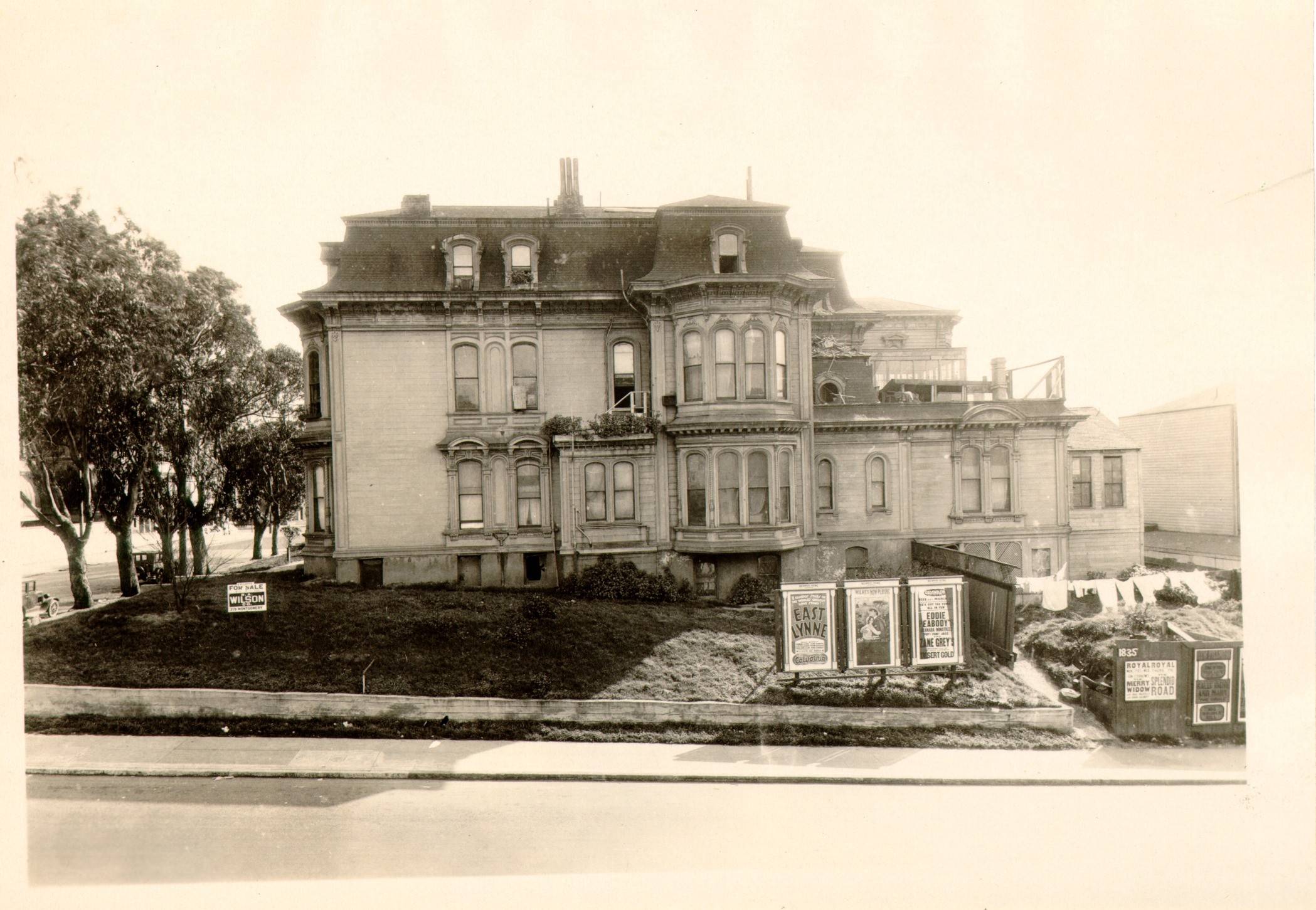 An old photograph of a large Victorian mansion on a grassy rise above a street, with billboards and a For Sale sign on the lot and a row of tall eucalyptus trees at the left