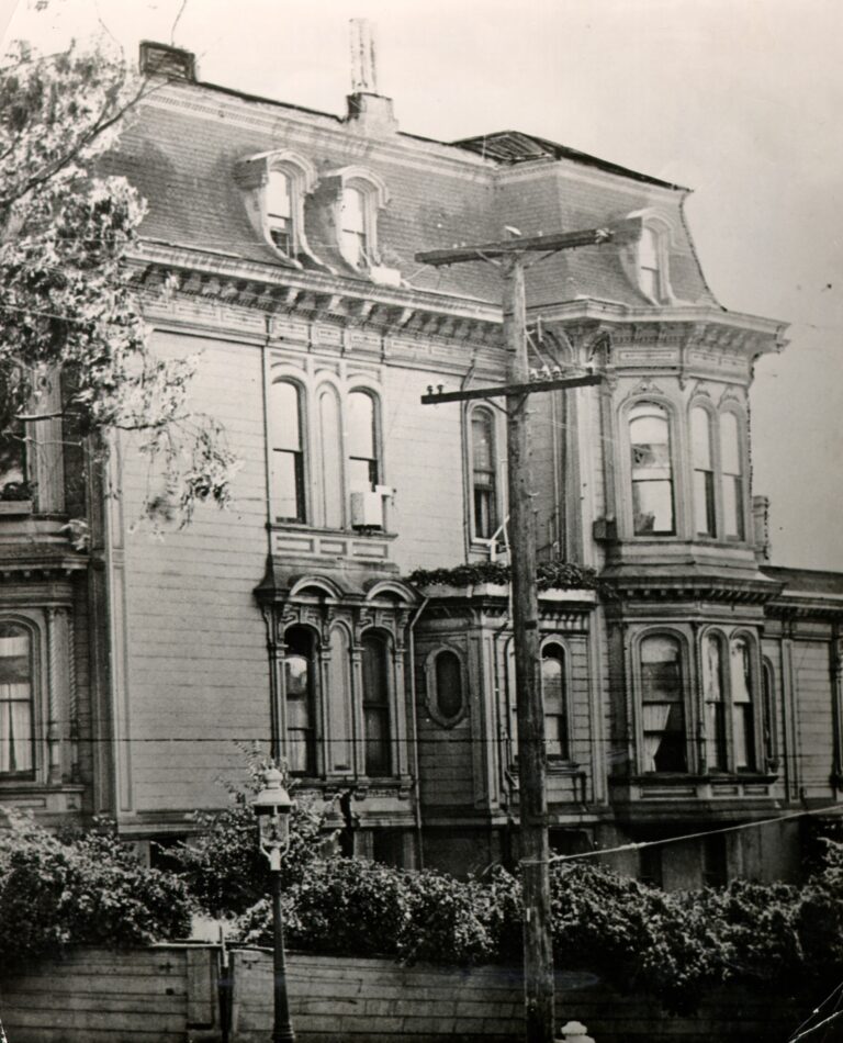 An old photograph of the upper floors of a grey Victorian mansion with a mansard roof, arched windows and bay windows, behind a wooden fence and hedge, with a telephone pole in front