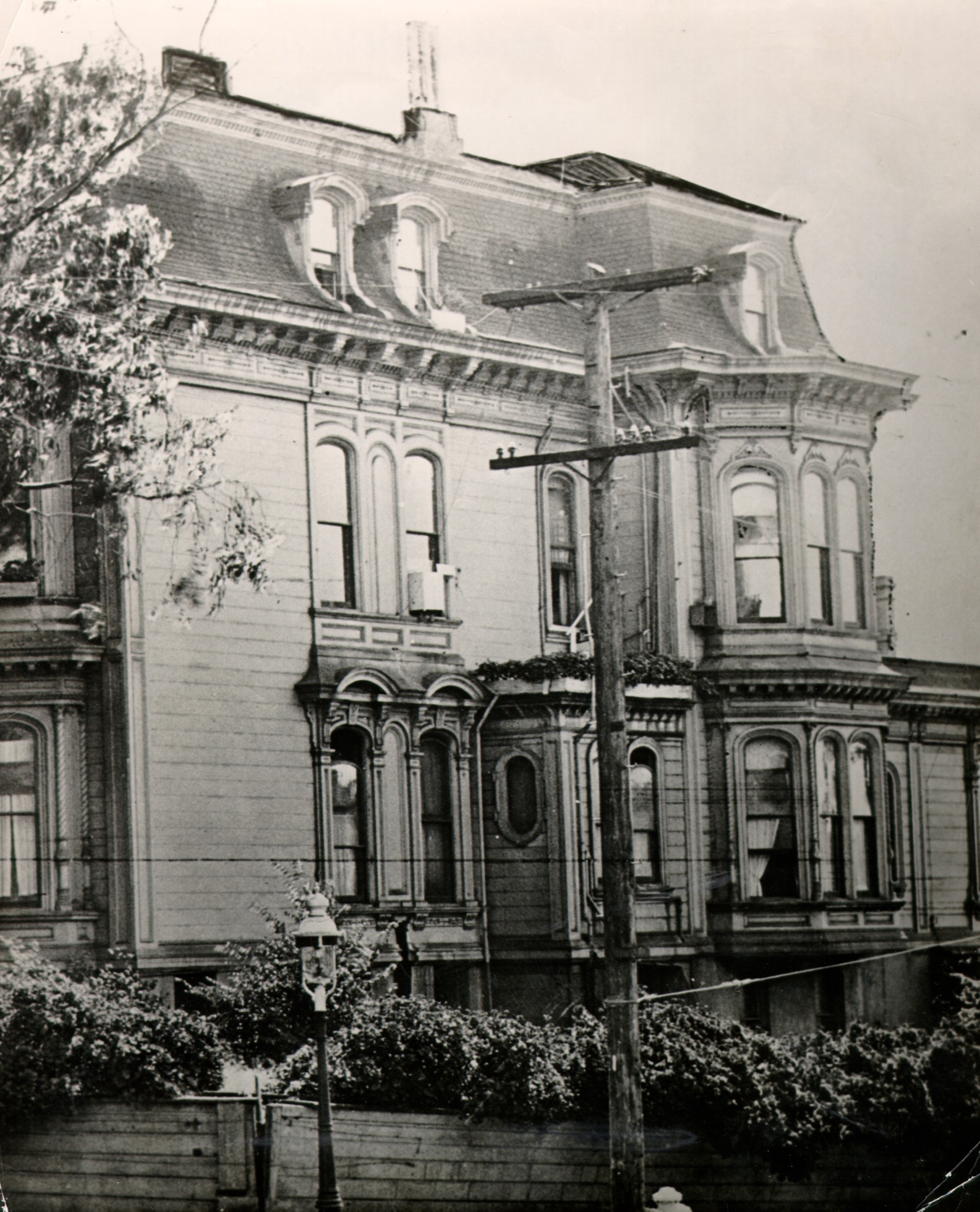 An old photograph of the upper floors of a grey Victorian mansion with a mansard roof, arched windows and bay windows, behind a wooden fence and hedge, with a telephone pole in front