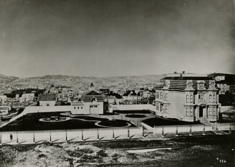 An old photograph of a large three-storey Victorian mansion with a mansard roof beside a walled formal garden of curving flower beds, seen across a bare lot, with the houses and hills of San Francisco behind