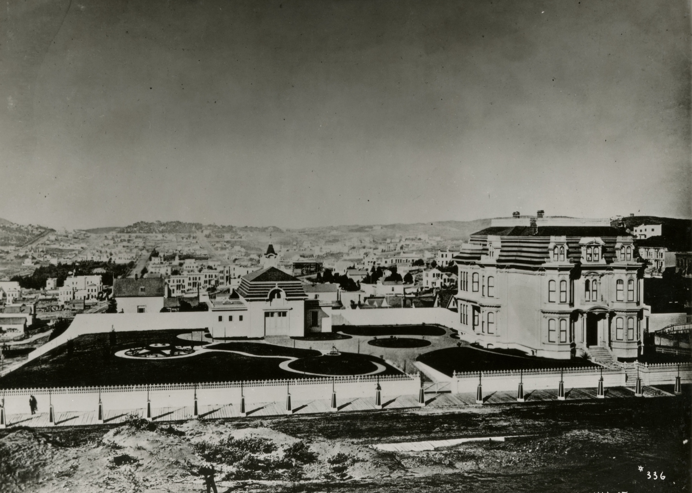 An old photograph of a large three-storey Victorian mansion with a mansard roof beside a walled formal garden of curving flower beds, seen across a bare lot, with the houses and hills of San Francisco behind