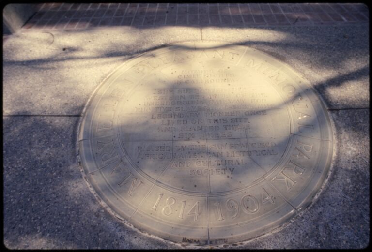 A round plaque set in a sidewalk, dappled with tree shadow, inscribed Mary Ellen Pleasant Memorial Park, 1814 to 1904