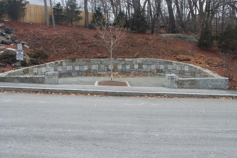 The Proctor's Ledge Memorial on Pope Street: a curved granite wall set with name stones, a young tree at its center and the wooded ledge rising behind