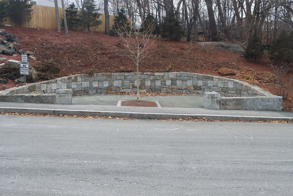 The Proctor's Ledge Memorial on Pope Street: a curved granite wall set with name stones, a young tree at its center and the wooded ledge rising behind