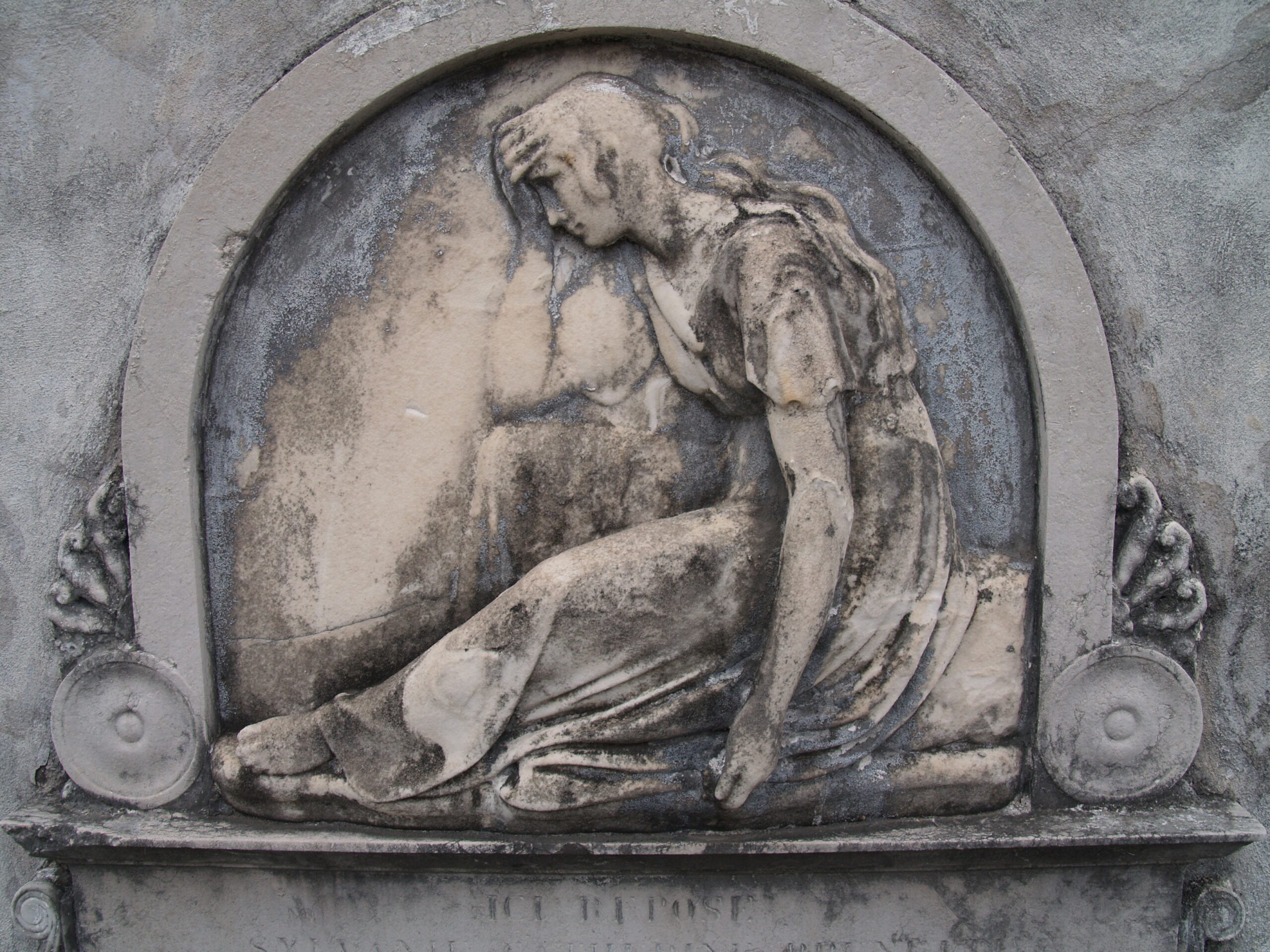 A stone relief of a seated woman bowing her head into her hand, inside an arch