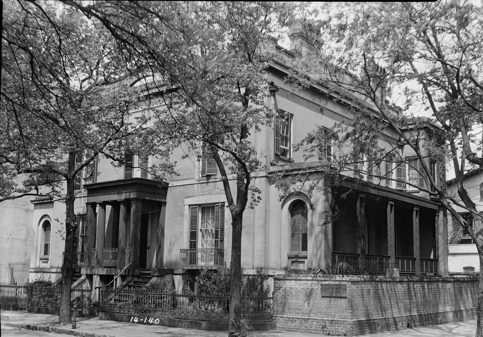 A black-and-white photo of a large two-story stucco house with a columned porch behind trees on a corner