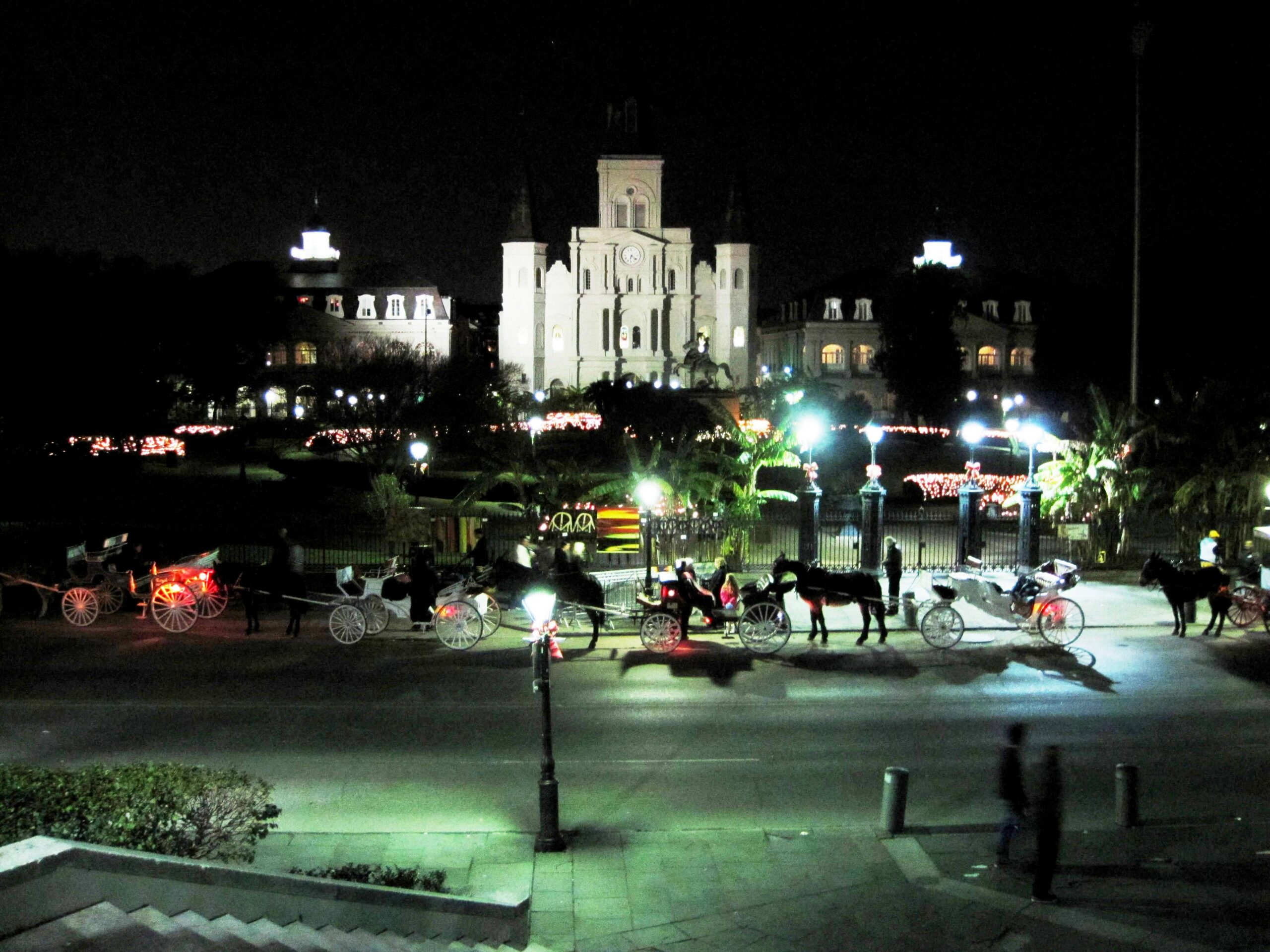 St. Louis Cathedral lit up at night above Jackson Square, with horse-drawn carriages lined up in the street