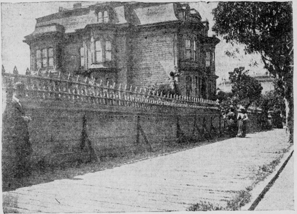 A grainy newspaper photograph of a three-storey Victorian mansion with a mansard roof, set back behind a long, high garden wall with an iron railing along a sloping sidewalk