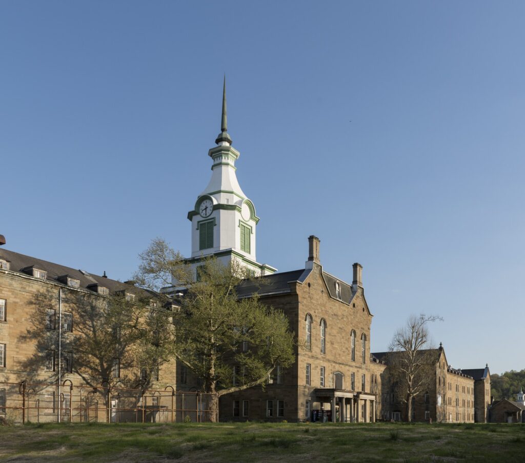 The sandstone central block of the asylum, its white clock tower against a clear blue sky, with long wings stretching to either side behind trees