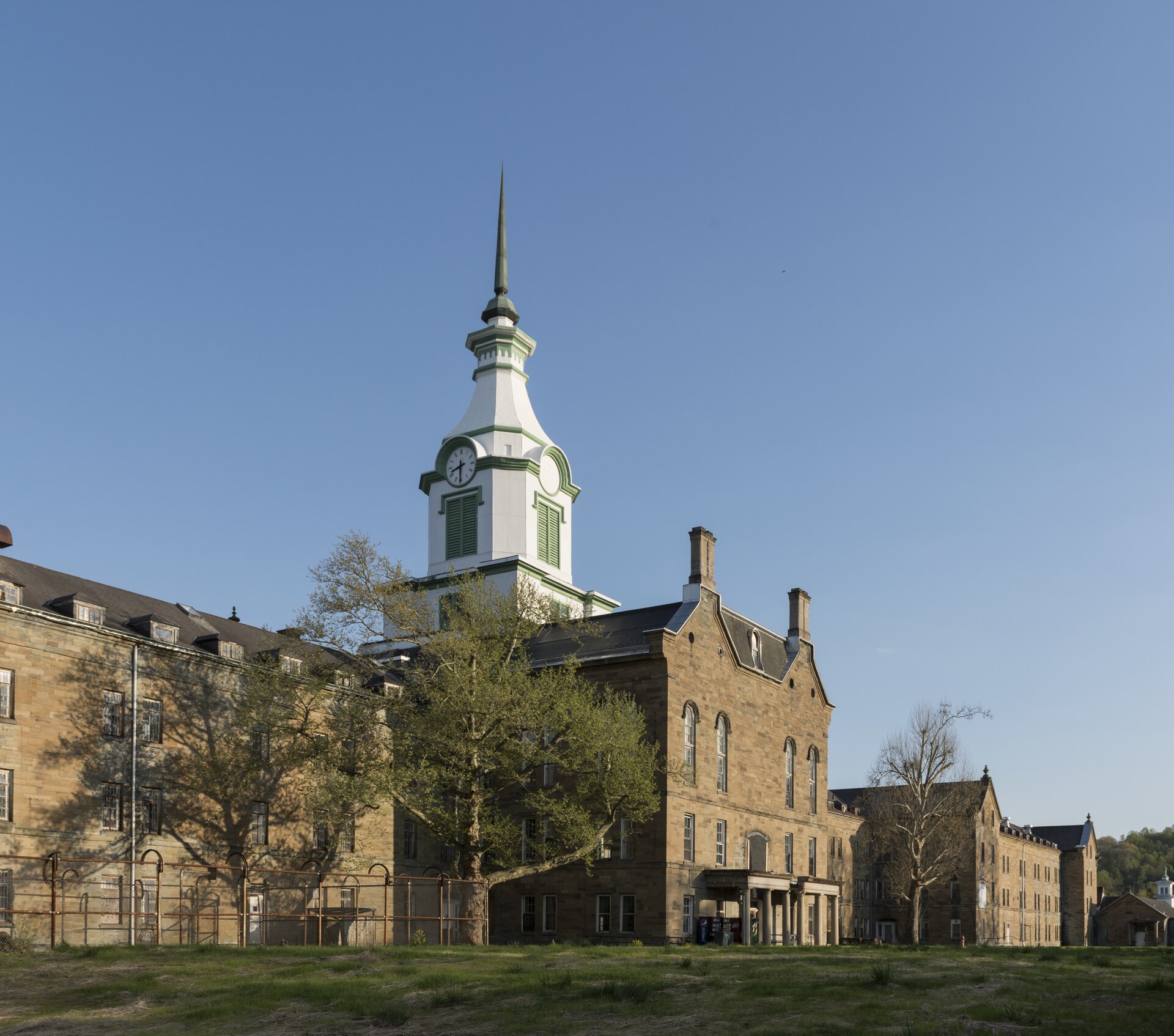 The sandstone central block of the asylum, its white clock tower against a clear blue sky, with long wings stretching to either side behind trees