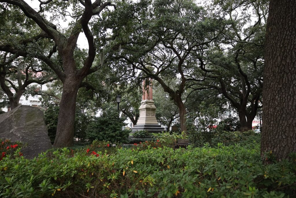 Live oaks over a square with a large boulder on the left and a tall stone monument among red flowers
