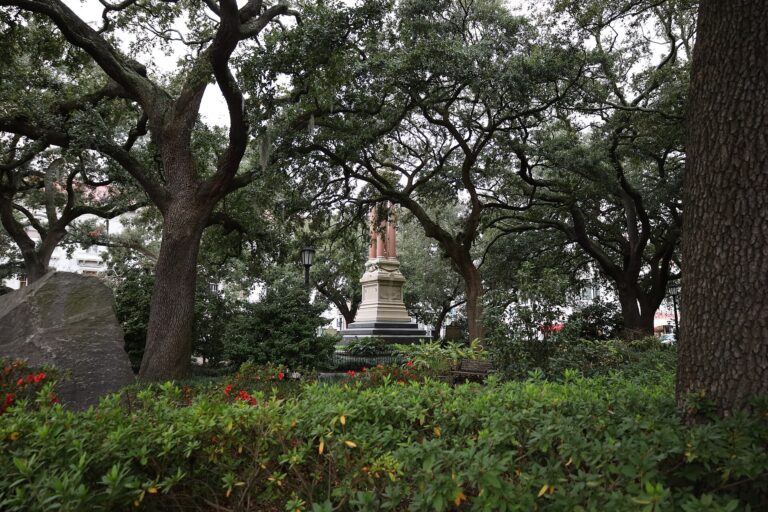 Live oaks over a square with a large boulder on the left and a tall stone monument among red flowers